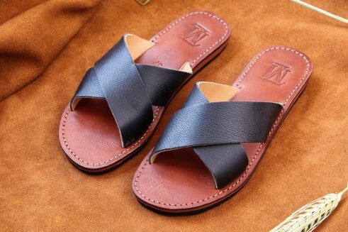 A pair of handmade Moroccan sandals with smooth black leather cross straps and natural brown leather soles, placed on a camel suede surface with wheat accents.