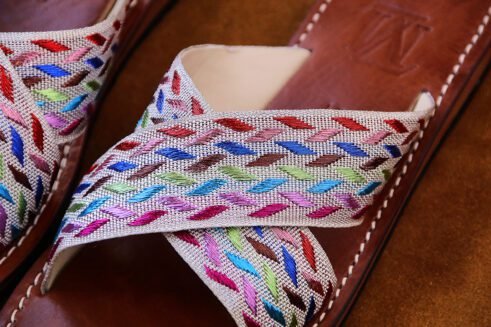 A pair of handmade Moroccan sandals with cross-over braided textile straps in multicolored threads on brown leather soles, placed on a camel suede background with wheat accents