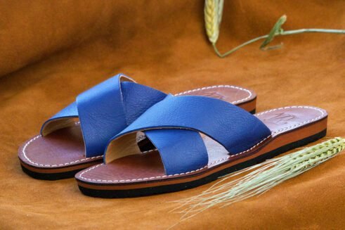 A pair of handmade Moroccan sandals featuring vibrant blue leather cross straps, brown stitched leather soles, and a natural beige leather lining, placed on a camel suede background with wheat stems.