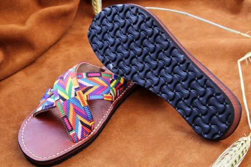 A pair of handmade Moroccan sandals featuring multicolor ethnic-patterned woven fabric in a cross-strap design over brown leather soles, displayed on a camel suede background with wheat accents