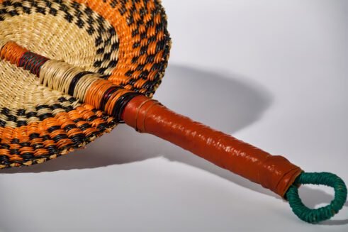 A handcrafted Moroccan fan with vibrant orange, black, and beige woven design, complemented by a leather handle, placed on a rocky surface with a scenic background