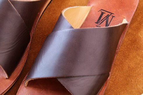 A pair of handmade Moroccan sandals featuring wide dark brown leather cross straps and stacked leather soles with a black rubber base, set on a camel suede background with wheat decor