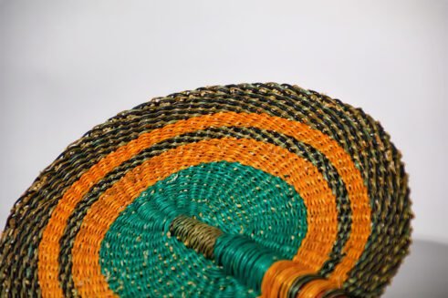 A handcrafted Moroccan fan featuring a vibrant green and orange circular design with a leather handle, placed on a rock with a blurred desert background.