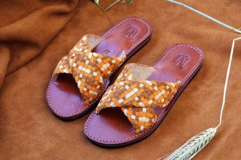 A pair of handmade Moroccan sandals with wide handwoven leather cross straps in beige and camel tones, brown leather soles, and white stitching, set on a camel suede background