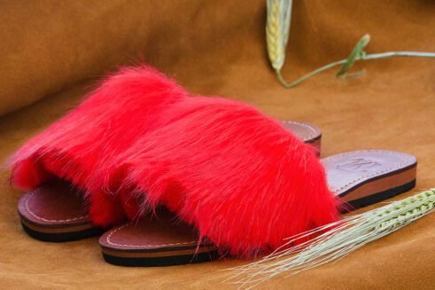 A pair of handmade Moroccan slide sandals featuring vibrant red hair-on leather uppers and brown leather soles, laid on a camel suede background with natural wheat details.