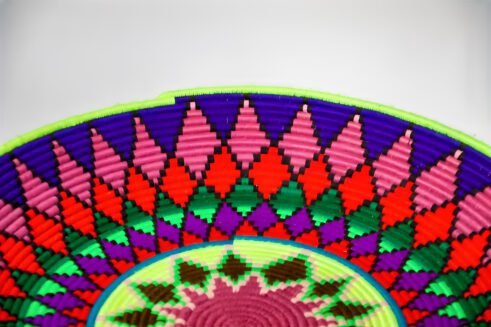 A handcrafted Moroccan coil basket with a vibrant geometric pattern in red, green, blue, purple, and yellow, placed on a neutral background with small stones surrounding it.