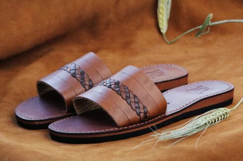 A pair of handmade Moroccan sandals in chestnut brown leather featuring a broad strap with a dark braided leather detail, brown stitched soles, and a black rubber base, styled on a suede background with wheat decoration