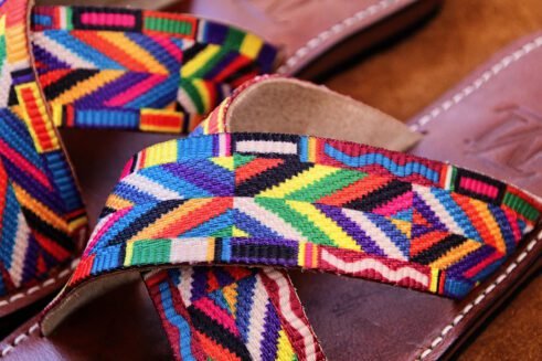 A pair of handmade Moroccan sandals featuring multicolor ethnic-patterned woven fabric in a cross-strap design over brown leather soles, displayed on a camel suede background with wheat accents