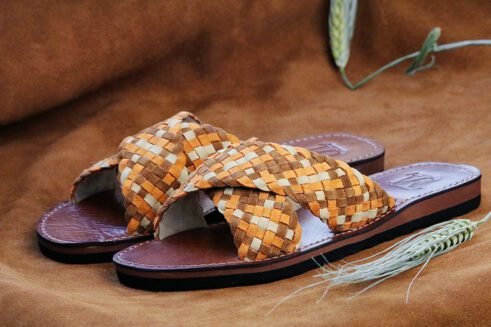 A pair of handmade Moroccan sandals with wide handwoven leather cross straps in beige and camel tones, brown leather soles, and white stitching, set on a camel suede background