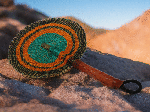 A handcrafted Moroccan fan featuring a vibrant green and orange circular design with a leather handle, placed on a rock with a blurred desert background.