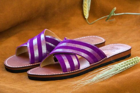 A pair of handmade Moroccan sandals with cross-over striped violet leather straps and brown leather soles, placed on a soft camel suede surface with wheat accents.