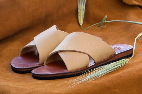 A pair of handmade Moroccan leather slide sandals with beige cross-over straps and brown leather soles, styled on a camel suede background with wheat accents.