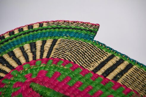 A handcrafted Moroccan fan featuring vibrant green, pink, yellow, and black colors with a leather handle, placed on a rocky surface with a scenic backdrop.