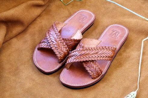 Pair of brown handwoven leather Moroccan sandals with crisscross braided straps, displayed on a warm suede background with barley spikes