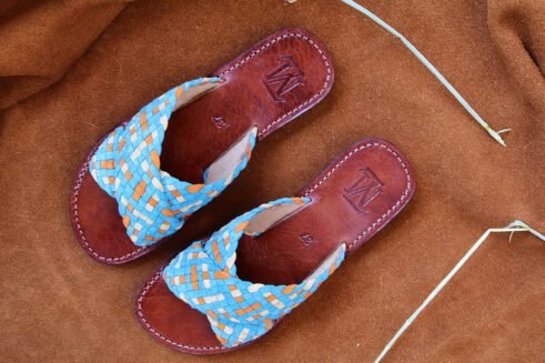 Handwoven leather sandals in blue, orange, and beige tones with a brown sole, placed on a suede background