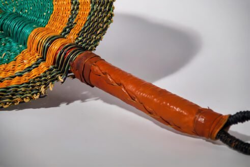 A handcrafted Moroccan fan featuring a vibrant green and orange circular design with a leather handle, placed on a rock with a blurred desert background.