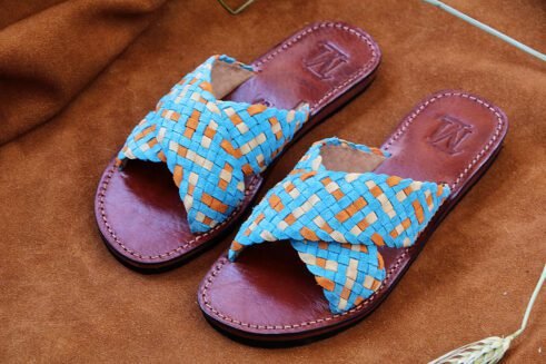 Handwoven leather sandals in blue, orange, and beige tones with a brown sole, placed on a suede background