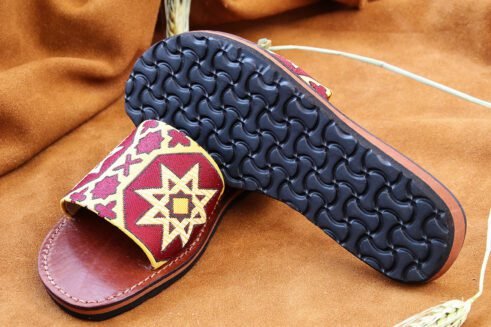 Pair of Moroccan leather sandals with embroidered red and gold star and geometric motifs inspired by traditional zellige tiles, photographed on a camel suede background with wheat stems