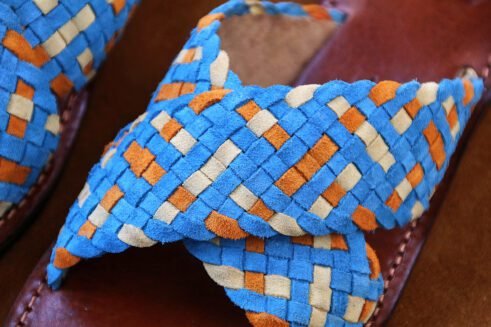 Handwoven leather sandals in blue, orange, and beige tones with a brown sole, placed on a suede background