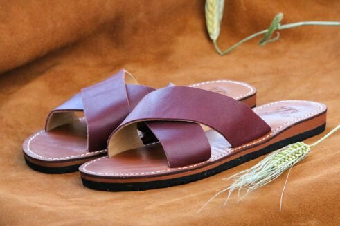A pair of handmade Moroccan sandals featuring rich plum-colored leather cross straps and brown leather soles with white stitching, styled on a camel suede background with wheat stems