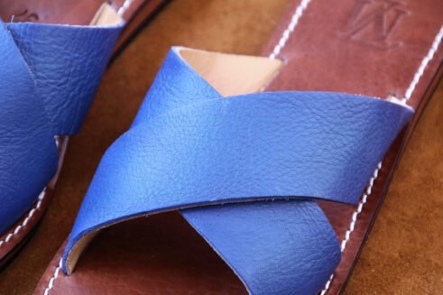 A pair of handmade Moroccan sandals featuring vibrant blue leather cross straps, brown stitched leather soles, and a natural beige leather lining, placed on a camel suede background with wheat stems.