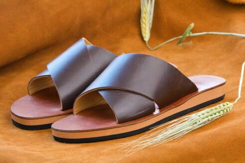 A pair of handmade Moroccan sandals featuring wide dark brown leather cross straps and stacked leather soles with a black rubber base, set on a camel suede background with wheat decor