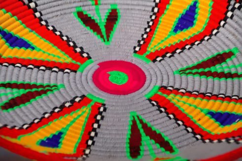 A beautifully handcrafted Moroccan coil basket with vibrant red, yellow, green, and blue geometric patterns, sitting on a white background surrounded by small stones.