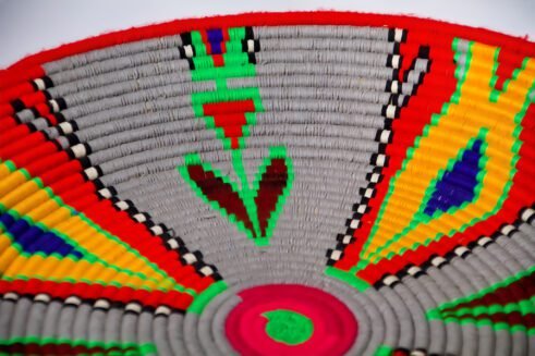 A beautifully handcrafted Moroccan coil basket with vibrant red, yellow, green, and blue geometric patterns, sitting on a white background surrounded by small stones.