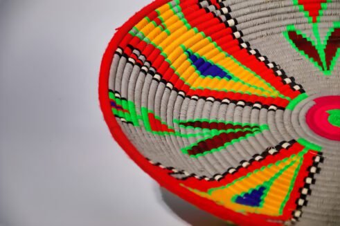 A beautifully handcrafted Moroccan coil basket with vibrant red, yellow, green, and blue geometric patterns, sitting on a white background surrounded by small stones.