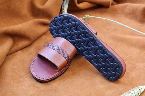 A pair of handmade Moroccan sandals in chestnut brown leather featuring a broad strap with a dark braided leather detail, brown stitched soles, and a black rubber base, styled on a suede background with wheat decoration