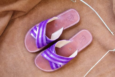 A pair of handmade Moroccan sandals with cross-over striped violet leather straps and brown leather soles, placed on a soft camel suede surface with wheat accents.
