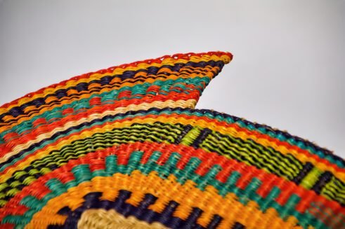A handcrafted Moroccan fan featuring a vibrant multicolor design with orange, blue, yellow, and red tones, with a leather handle, placed on a rocky surface with a mountain backdrop.