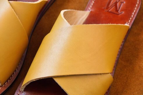 A pair of handmade Moroccan sandals featuring smooth ochre tan leather cross straps, brown leather footbeds, and black rubber soles, placed on a camel suede background with decorative wheat