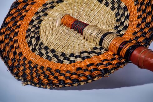 A handcrafted Moroccan fan with vibrant orange, black, and beige woven design, complemented by a leather handle, placed on a rocky surface with a scenic background