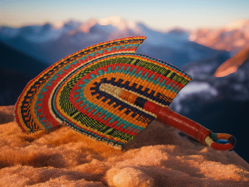 A handcrafted Moroccan fan featuring a vibrant multicolor design with orange, blue, yellow, and red tones, with a leather handle, placed on a rocky surface with a mountain backdrop.