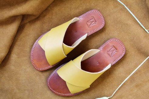 A pair of handmade Moroccan sandals featuring smooth ochre tan leather cross straps, brown leather footbeds, and black rubber soles, placed on a camel suede background with decorative wheat