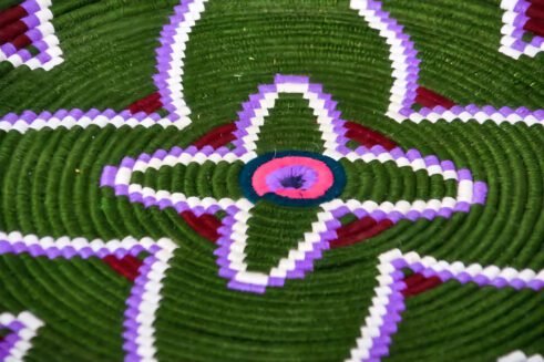A handcrafted Moroccan coil basket with a green and purple floral design, surrounded by small stones on a neutral background.