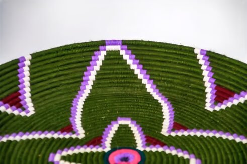 A handcrafted Moroccan coil basket with a green and purple floral design, surrounded by small stones on a neutral background.