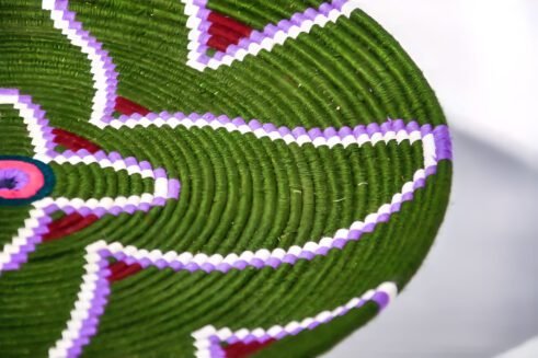 A handcrafted Moroccan coil basket with a green and purple floral design, surrounded by small stones on a neutral background.
