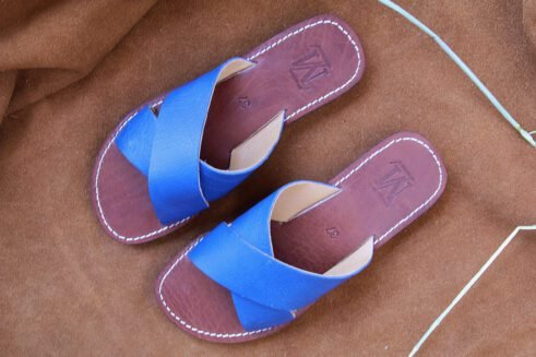 A pair of handmade Moroccan sandals featuring vibrant blue leather cross straps, brown stitched leather soles, and a natural beige leather lining, placed on a camel suede background with wheat stems.