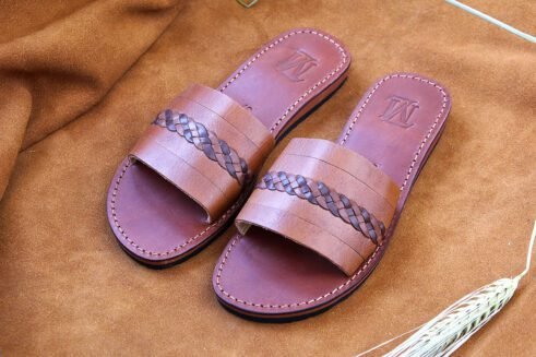 A pair of handmade Moroccan sandals in chestnut brown leather featuring a broad strap with a dark braided leather detail, brown stitched soles, and a black rubber base, styled on a suede background with wheat decoration