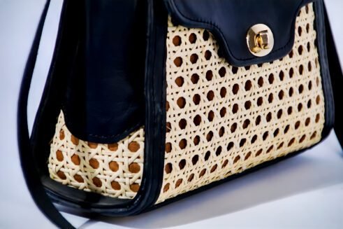 Chic handbag with a black leather top and woven cane body, featuring a gold-tone clasp, a sturdy handle, and an adjustable shoulder strap, styled against a soft neutral backdrop.