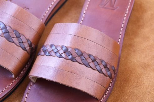 A pair of handmade Moroccan sandals in chestnut brown leather featuring a broad strap with a dark braided leather detail, brown stitched soles, and a black rubber base, styled on a suede background with wheat decoration