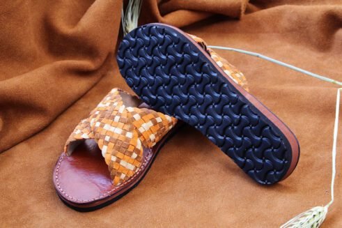 A pair of handmade Moroccan sandals with wide handwoven leather cross straps in beige and camel tones, brown leather soles, and white stitching, set on a camel suede background