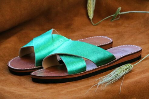 A pair of handmade Moroccan slide sandals with metallic emerald green cross-over leather straps, brown leather soles, and visible white stitching, displayed on a camel suede background.