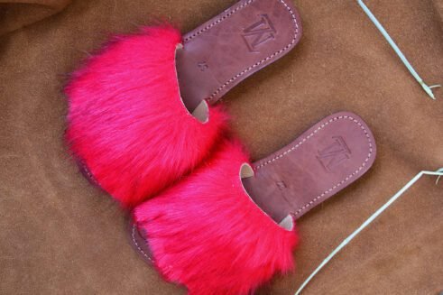 A pair of handmade Moroccan slide sandals featuring vibrant red hair-on leather uppers and brown leather soles, laid on a camel suede background with natural wheat details.