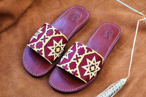 Pair of Moroccan leather sandals with embroidered red and gold star and geometric motifs inspired by traditional zellige tiles, photographed on a camel suede background with wheat stems