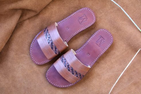 A pair of handmade Moroccan sandals in chestnut brown leather featuring a broad strap with a dark braided leather detail, brown stitched soles, and a black rubber base, styled on a suede background with wheat decoration