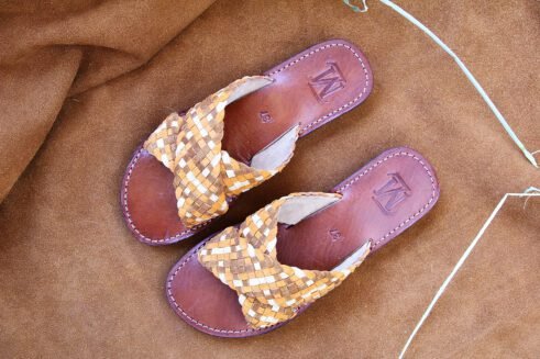A pair of handmade Moroccan sandals with wide handwoven leather cross straps in beige and camel tones, brown leather soles, and white stitching, set on a camel suede background