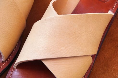 A pair of handmade Moroccan leather slide sandals with beige cross-over straps and brown leather soles, styled on a camel suede background with wheat accents.
